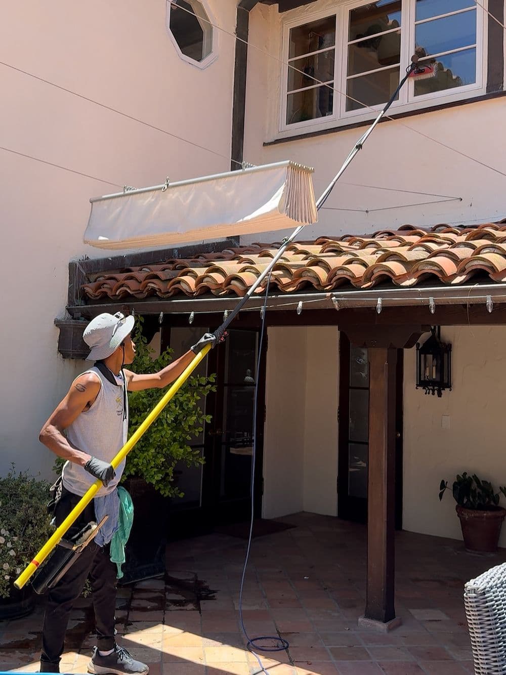 Man using a pole to clean windows on a house with a tiled roof and outdoor patio.