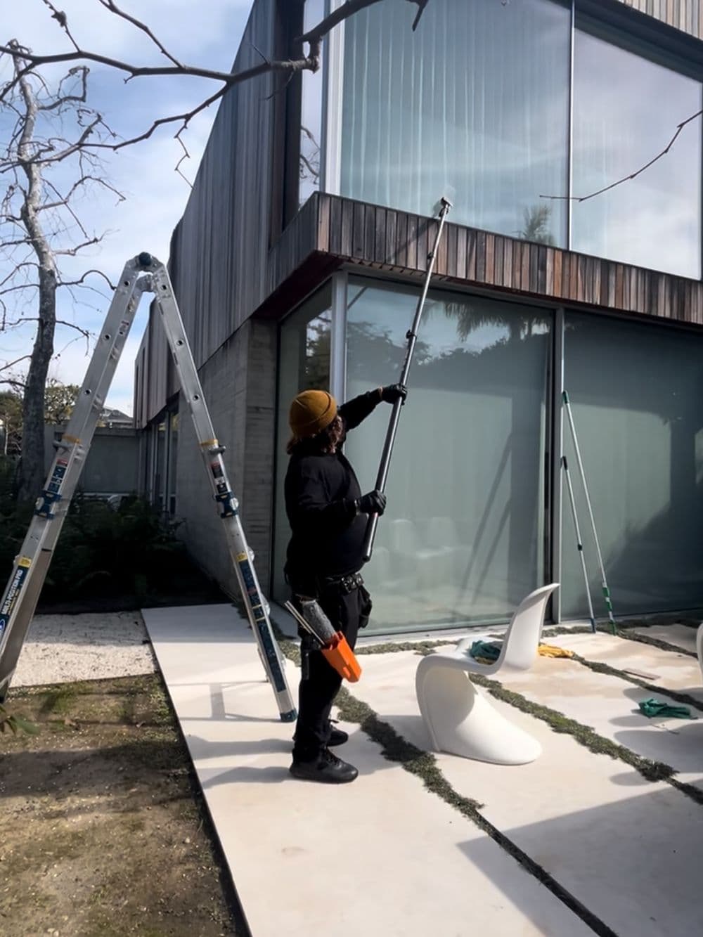 Person using a long pole to clean windows of a modern building with a ladder nearby.