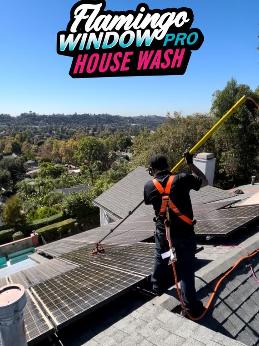 Person washing solar panels on a roof with a long pole, overlooking green hills.